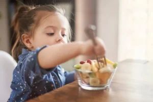 Child eating a fruit salad with a fork at a table.