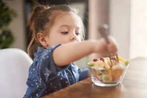 Young child eating a fruit salad with a fork at a wooden table.