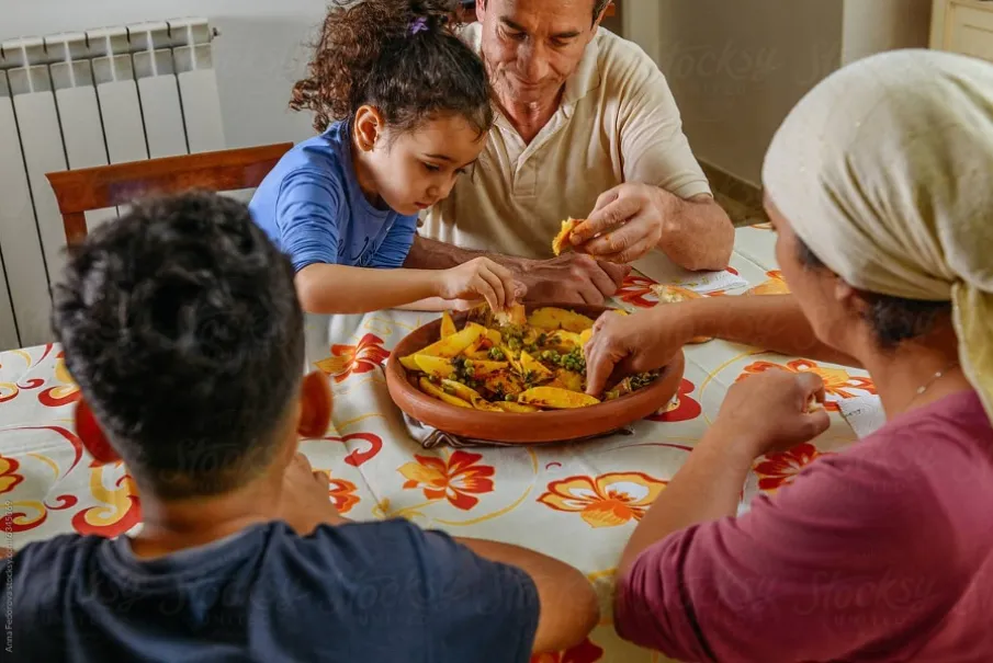Family with young child sharing a traditional dish indoors with colorful tablecloth.